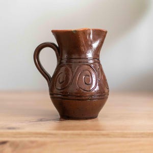 以下が含まれることがあります： 渦巻き模様の小さな茶色の陶器のピッチャー。ピッチャーは持ち手と広い口が付いています。