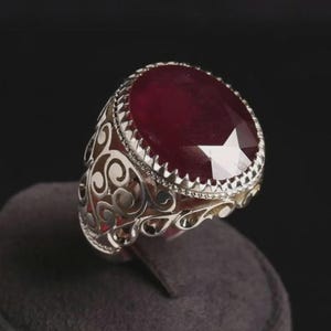 May include: A silver ring with an oval, faceted, deep red gemstone. The ring has an ornate, openwork design on the band. The gemstone is set in a serrated bezel.