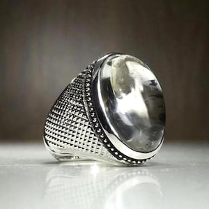 Pode incluir: Um anel de prata com uma pedra grande, oval e transparente. O anel tem uma banda texturizada com um padrão geométrico e uma borda com contas. A pedra é definida em uma moldura de prata polida.