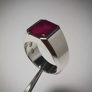May include: A silver ring with a large, rectangular, deep red gemstone. The ring has a polished finish and a classic, masculine design. The gemstone is set in a bezel setting, and the ring appears to be held by a pair of tweezers.
