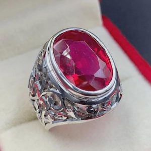 May include: A silver ring featuring an oval, faceted ruby gemstone. The ring has an ornate, filigree design on the band. The ruby is a deep, vibrant red color, and the ring is set against a soft, off-white background.