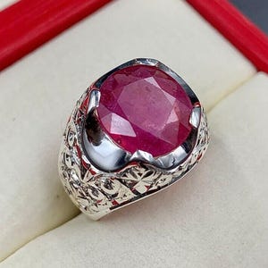 Può includere: Un anello d'argento con una grande gemma di rubino taglio ovale. Il rubino è di colore rosso intenso ed è incastonato in una fascia d'argento incisa e dettagliata. L'anello è esposto in un cofanetto rosso e bianco.