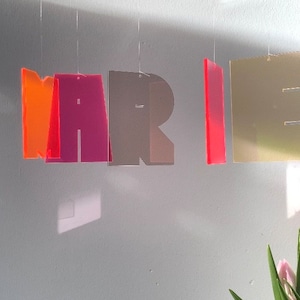 May include: A colourful mobile spelling out the name "MARIE" with translucent letters in orange, pink, grey, red, and gold.