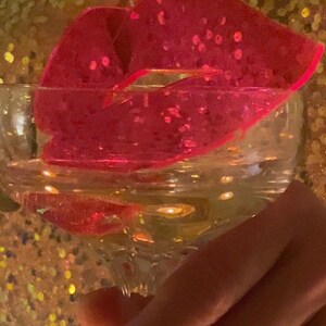 Pu&ograve; includere: Uno stampo per ghiaccio a forma di labbra rosa con glitter in un bicchiere trasparente pieno d'acqua.