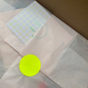 Pu&ograve; includere: Una scatola piena di carta velina bianca con un adesivo circolare giallo e un biglietto bianco con un motivo a griglia dorata.