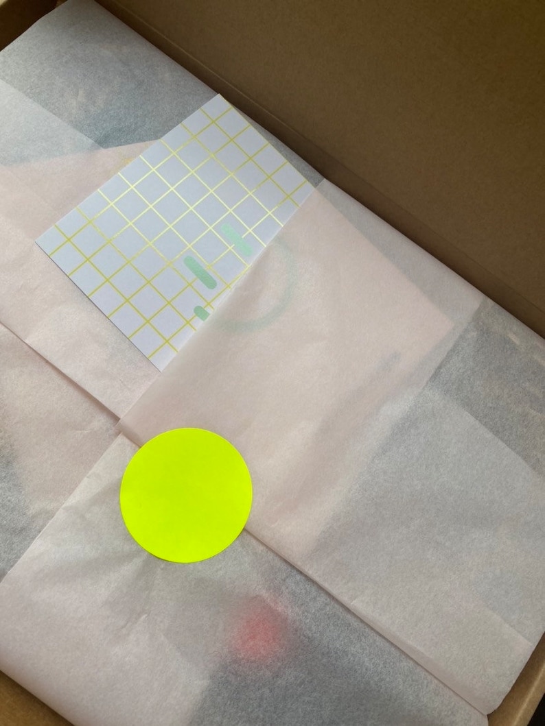 Pu&ograve; includere: Una scatola piena di carta velina bianca con un adesivo circolare giallo e un biglietto bianco con un motivo a griglia dorata.