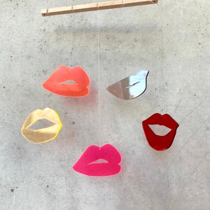 Pu&ograve; includere: Un mobile con cinque forme sospese, tra cui quattro labbra di diversi colori e un uccello argentato. Le labbra sono rosse, rosa, arancioni e gialle. Il mobile &egrave; sospeso da un'asta di legno.