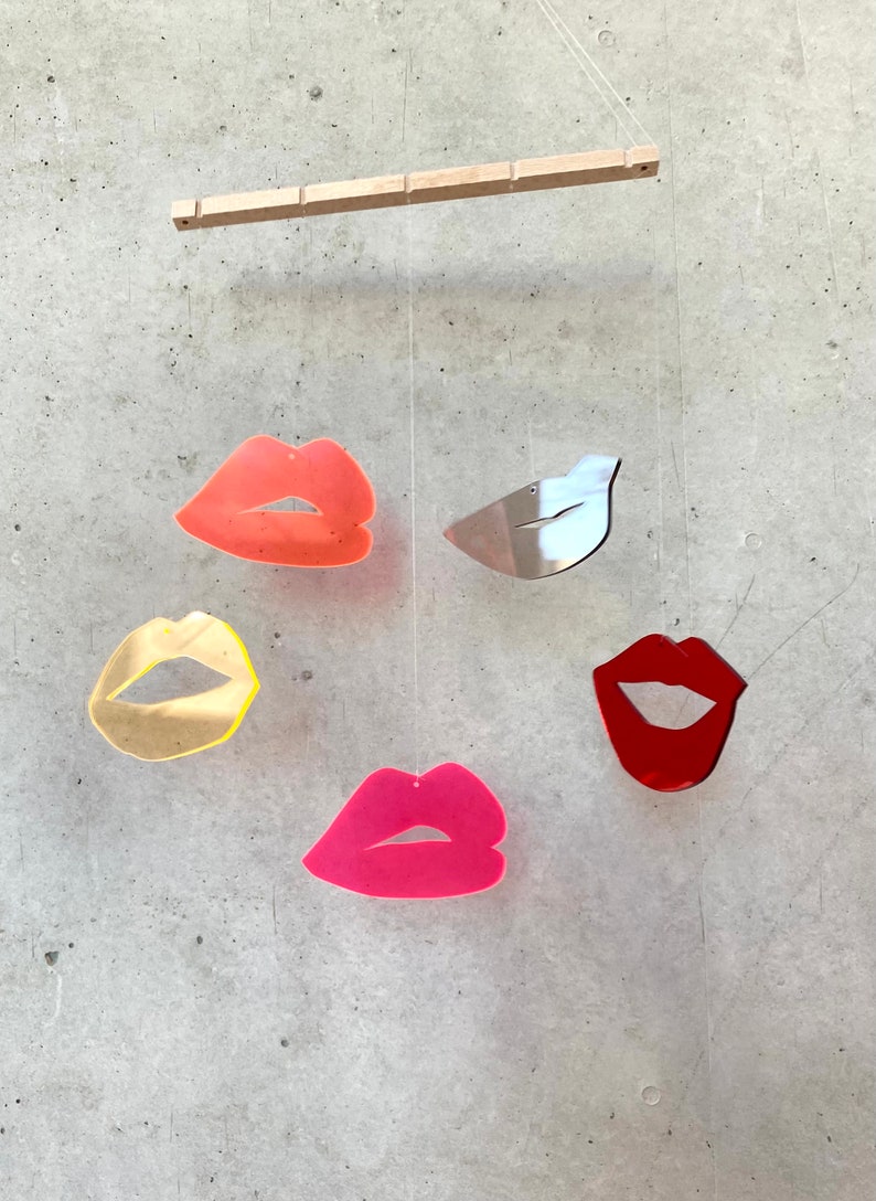 Pu&ograve; includere: Un mobile con cinque forme sospese, tra cui quattro labbra di diversi colori e un uccello argentato. Le labbra sono rosse, rosa, arancioni e gialle. Il mobile &egrave; sospeso da un'asta di legno.