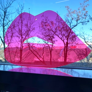 Könnte beinhalten: Ein rosa Plastik-Ausschnitt in Form von Lippen mit einem Loch in der Mitte. Der Ausschnitt ist auf einem Fenster mit Blick auf Bäume und einen blauen Himmel platziert.