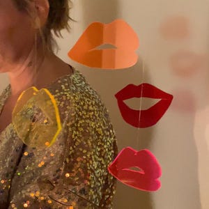 Pu&ograve; includere: Un set di quattro labbra di carta colorate appese a un soffitto. Le labbra sono in tonalit&agrave; di arancione, rosso e rosa.
