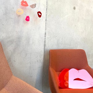 Pu&ograve; includere: Due sedie arancioni con un oggetto a forma di labbra rosa e rosse sul sedile di una delle sedie. Un mobile con diverse labbra colorate &egrave; appeso al soffitto sopra le sedie.