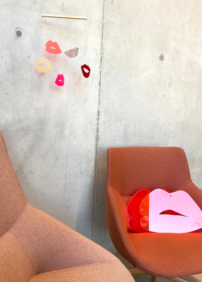 Pu&ograve; includere: Due sedie arancioni con un oggetto a forma di labbra rosa e rosse sul sedile di una delle sedie. Un mobile con diverse labbra colorate &egrave; appeso al soffitto sopra le sedie.