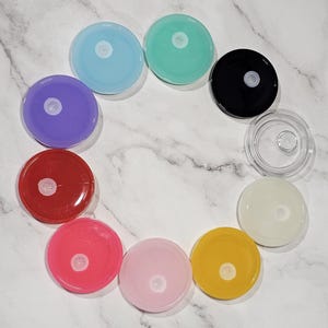 Peut inclure: Assortiment de dix couvercles ronds, plats et en plastique de différentes couleurs, dont le violet, le rouge, le rose, le bleu clair, le jaune, le blanc, le noir et le vert. Les couvercles ont un petit trou central en plastique transparent.