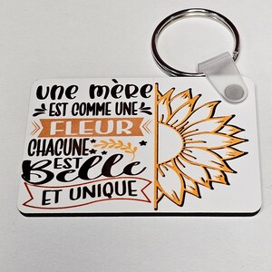 Peut inclure: Un porte-clés blanc avec un motif de tournesol et le texte "une mère est comme une fleur chacune est belle et unique".