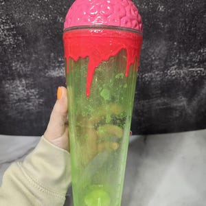 May include: A clear plastic tumbler with a pink brain-shaped lid and red dripping accents. The cup transitions from a green base to a clear body. The design is a novelty item.