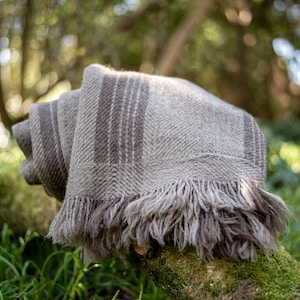 Op de afbeelding: Een gevouwen deken in neutrale tinten, met een visgraatpatroon en gefranjerde randen. De deken heeft verticale strepen in een donkerdere bruine tint. De zachte, getextureerde plaid wordt buiten gepresenteerd, rustend op een met mos bedekte boomstam.