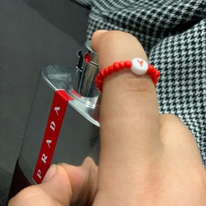 Könnte beinhalten: Ein roter Perlenring mit einer weißen Perle mit dem Buchstaben "Y". Der Ring befindet sich an einem Finger neben einer silbernen und roten Prada-Parfümflasche. Der Hintergrund enthält ein schwarz-weißes Hahnentrittmuster.