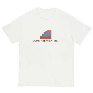 Puede incluir: Una camiseta blanca de manga corta con un gráfico de cinco lomos de libros rectangulares apilados en naranja, rosa, turquesa y rojo, con el texto "A COURT OF THORNS AND ROSES" y "26,000 STEPS LATER..." impreso en letras negras.