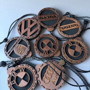 May include: A selection of wooden car logo ornaments, including Volkswagen, Volvo, Porsche, BMW, and Audi. Each ornament features a laser-cut logo within a circular frame, with a dark grey backing and a black cord for hanging. The wood has a warm, reddish-brown tone.