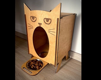 Puede incluir: Un comedero para mascotas de madera con forma de gato con un cuenco elevado y una bandeja extraíble. El comedero tiene un recorte en forma de cara de gato con una expresión de sorpresa.