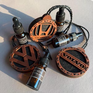 Op de afbeelding: Drie ronde houten auto-luchtverfrissers met de logo's van Honda, Volkswagen en Volvo, elk voorzien van een zwart gevlochten koord. Drie kleine amberkleurige glazen druppelflesjes met zwarte doppen en etiketten liggen tussen de houten ornamenten.