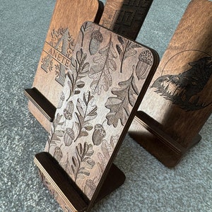 May include: Three wooden phone stands with different engraved designs. The first stand has a leafy pattern with acorns, the second has a geometric pattern, and the third has a wolf howling at the moon.
