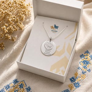Op de afbeelding: Een zilveren hangerketting met een ronde bedel met een kaartomtrek en tekst in een witte geschenkdoos. De ketting wordt gepresenteerd op een achtergrond van stof met patronen. Het merklogo is zichtbaar.