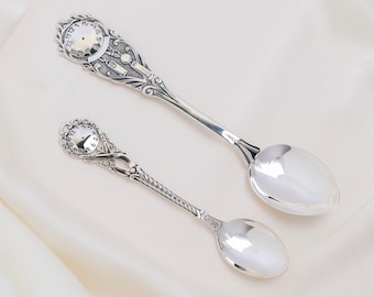 May include: Two silver spoons with ornate clock-face designs on the handles. The spoons have a polished finish and are set against a white background. The larger spoon is approximately 7 inches long, and the smaller spoon is about 5 inches long.