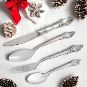 May include: Four silver-plated utensils with ornate handles, including a knife, fork, and two spoons, arranged on a white surface.