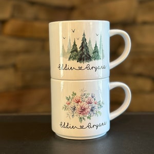 May include: Two white ceramic mugs stacked on top of each other. The top mug has a watercolor design of green trees and the text "Eddie & Aryana". The bottom mug has a watercolor design of pink and blue flowers and the text "Eddie & Aryana".