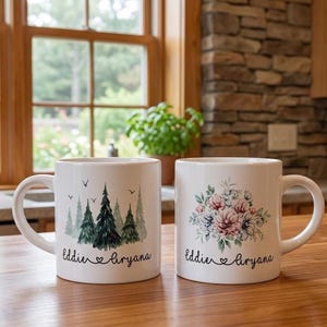 May include: Two white ceramic mugs with handles. The left mug features a watercolor forest of green pine trees and birds with the text Eddie and Liryana. The right mug displays a floral bouquet of pink and blue flowers with the text Eddie and Liryana.