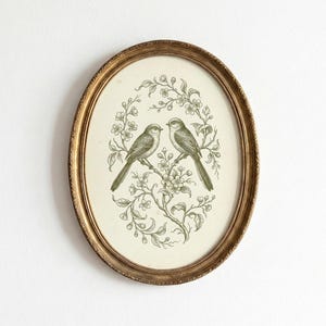 Peut inclure: Impression ovale encadrée avec deux oiseaux sur une branche fleurie. Les oiseaux et le motif floral sont dans des tons verts sur un fond crème. Le cadre est doré avec une bordure texturée. Décoration murale de style vintage.