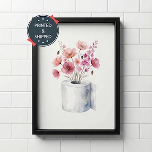 May include: A framed wall art print featuring a watercolor illustration of a white toilet paper roll serving as a vase for a bouquet of pink and red flowers. A circular badge in the top left corner contains the text PRINTED & SHIPPED.