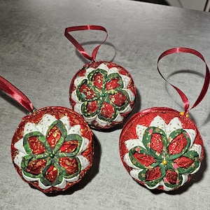 May include: Three handmade Christmas baubles. Each bauble is a red sphere with a floral design in green, white, and gold. A red ribbon is attached for hanging. The baubles are on a grey surface.