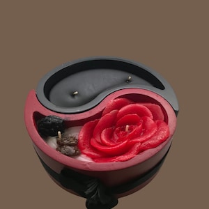 Può includere: Una candela yin yang rossa e nera con una rosa rossa e bacche nere in cima. La candela ha la forma di un simbolo yin yang.