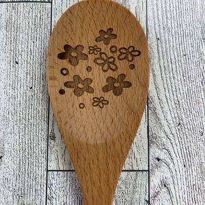 Op de afbeelding: Een houten lepel met een bloemmotief dat in de voorkant is gegraveerd. De lepel is lichtbruin en heeft een natuurlijke houten afwerking.