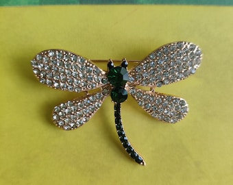 May include: A gold-coloured dragonfly brooch with clear and emerald green crystals. The wings are encrusted with clear crystals, while the body and tail feature green crystals. The brooch is a decorative accessory.