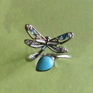 May include: A silver ring with a dragonfly design featuring blue and white enamel and clear rhinestones. The ring has a turquoise stone in the centre.