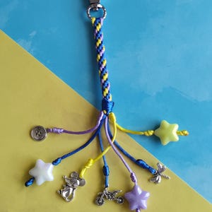 May include: A blue, yellow, and purple braided keychain with a silver clip. The keychain has several charms, including a star, a mouse, a button, a dragonfly, and a motorcycle.