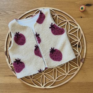 May include: A sleeveless vest made of cream-colored sherpa fleece fabric featuring a repeating pattern of dark magenta strawberry shapes with small dark brown stems. The vest has a simple open front design and a smooth white interior lining.