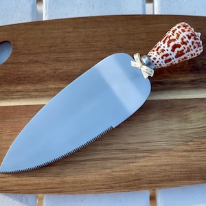 May include: A wooden cutting board with a cake server. The server has a silver blade with a serrated edge and a handle made from a seashell with brown and white spots. A small bow is attached to the server.