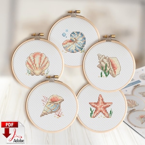 Puede incluir: Cinco bastidores de bordado circulares de madera con tela Aida blanca y patrones de punto de cruz de varias conchas marinas y una estrella de mar. Los diseños incluyen hilos de colores coral, rosa, azul y tostado, mostrando un nautilo y caracolas.