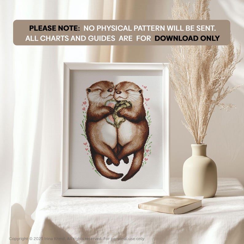 May include: A digital cross-stitch pattern featuring two otters cuddling. The otters are brown and white, and they are surrounded by pink flowers. The pattern is in a white frame and is sitting on a table.