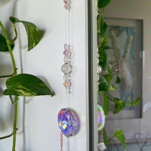 May include: An iridescent crystal suncatcher with a faceted pendant and crystal beads. The suncatcher hangs from a thin string, creating a rainbow effect. The design includes a chain and smaller crystal accents. The suncatcher is a decorative item.