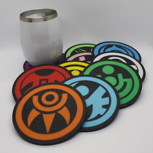 Op de afbeelding: Een verzameling kleurrijke, ronde onderzetters met diverse geometrische ontwerpen in oranje, blauw, groen en andere kleuren. Een roestvrijstalen beker staat naast de onderzetters. De onderzetters hebben een zwarte rand.
