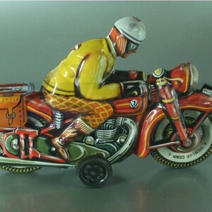 K&ouml;nnte beinhalten: Ein rotes und gelbes Spielzeugmotorrad mit einem Fahrer, der einen wei&szlig;en Helm und eine gelbe Jacke tr&auml;gt. Das Motorrad hat eine braune Satteltasche und die Worte "DUNLOP CORD" sind auf dem Hinterreifen aufgedruckt.