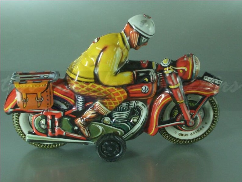 K&ouml;nnte beinhalten: Ein rotes und gelbes Spielzeugmotorrad mit einem Fahrer, der einen wei&szlig;en Helm und eine gelbe Jacke tr&auml;gt. Das Motorrad hat eine braune Satteltasche und die Worte "DUNLOP CORD" sind auf dem Hinterreifen aufgedruckt.