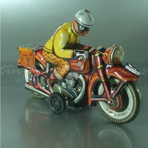 K&ouml;nnte beinhalten: Ein rotes und gelbes Spielzeugmotorrad mit einem Fahrer, der einen wei&szlig;en Helm und eine gelbe Jacke tr&auml;gt. Das Motorrad hat eine braune Ledertasche und einen schwarz-wei&szlig;en Reifen mit dem Wort "Gothard" darauf.