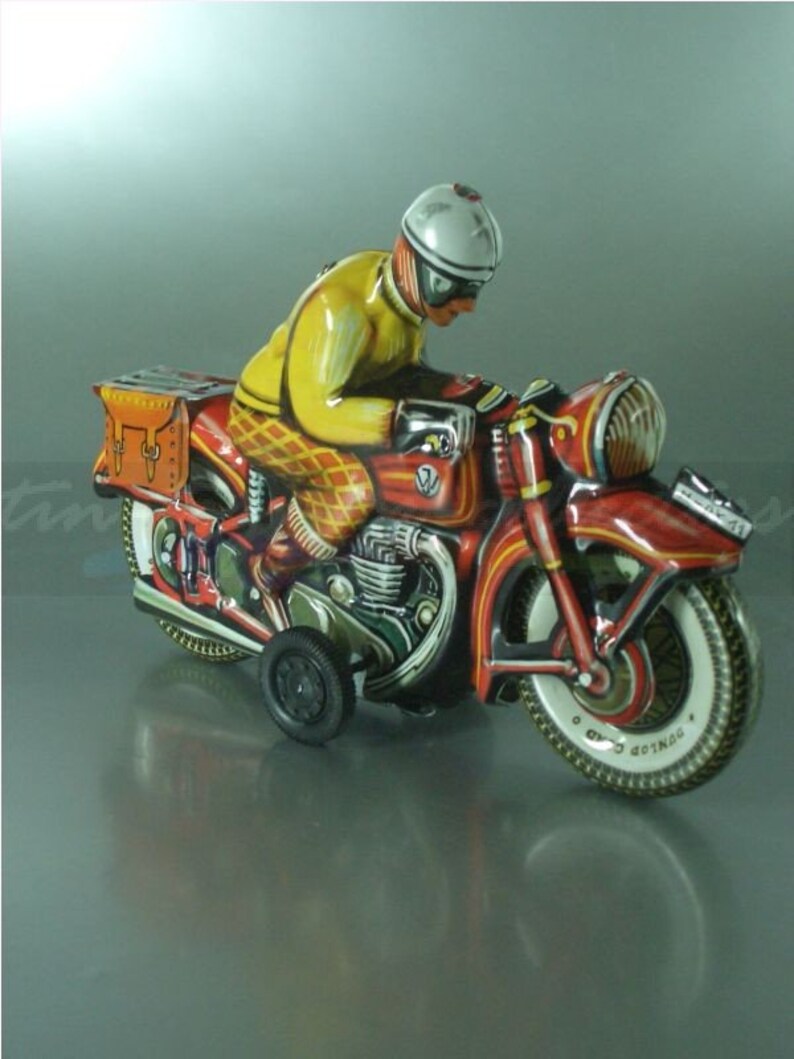 K&ouml;nnte beinhalten: Ein rotes und gelbes Spielzeugmotorrad mit einem Fahrer, der einen wei&szlig;en Helm und eine gelbe Jacke tr&auml;gt. Das Motorrad hat eine braune Ledertasche und einen schwarz-wei&szlig;en Reifen mit dem Wort "Gothard" darauf.