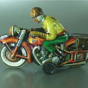 K&ouml;nnte beinhalten: Ein rotes, gelbes und gr&uuml;nes Spielzeugmotorrad mit einem Fahrer, der einen braunen Helm und eine Schutzbrille tr&auml;gt. Das Motorrad hat einen schwarzen Beiwagen mit einer braunen Tasche im Inneren. Das Motorrad hat einen schwarzen Dunlop-Reifen am Vorderrad und einen schwarzen Reifen am Hinterrad. Das Motorrad hat ein schwarzes Nummernschild mit der Aufschrift "N-HO 114".